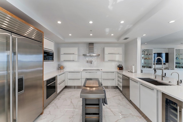 kitchen with built in appliances, wall chimney exhaust hood, white cabinetry, and sink