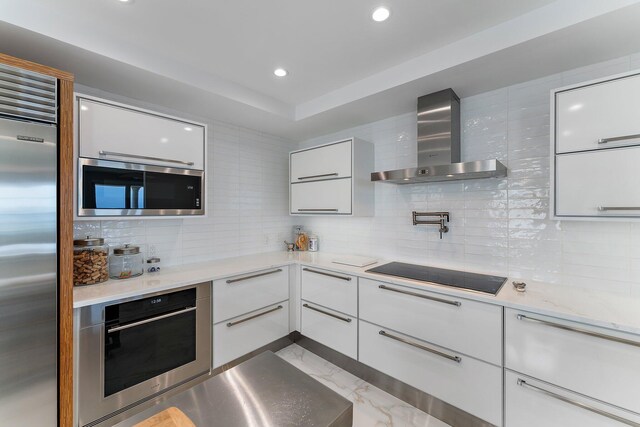 kitchen featuring light stone countertops, sink, wall chimney range hood, built in appliances, and white cabinets