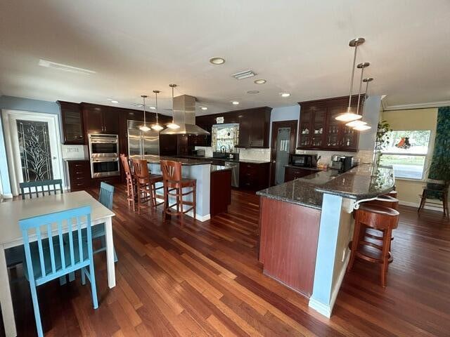 kitchen with a kitchen island, dark brown cabinets, dark wood-type flooring, exhaust hood, and appliances with stainless steel finishes