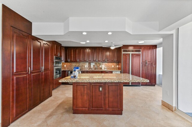 kitchen featuring ceiling fan, oven, light stone counters, a kitchen island, and backsplash