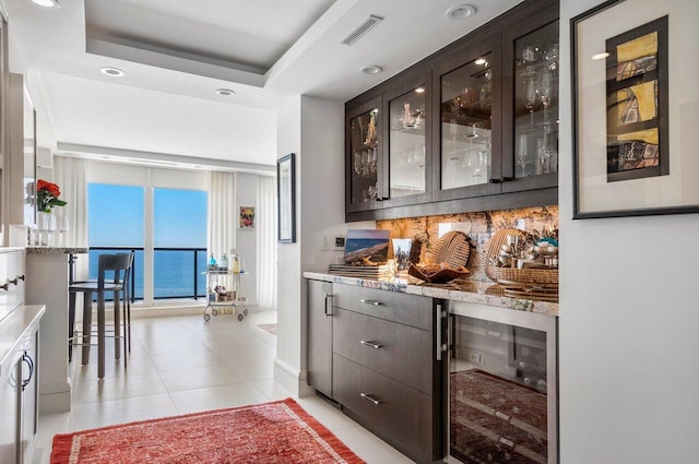 bar with light tile patterned flooring, wine cooler, dark brown cabinetry, a tray ceiling, and light stone countertops