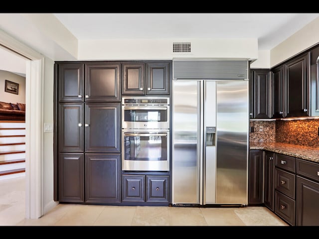 kitchen with dark brown cabinets, light tile patterned floors, appliances with stainless steel finishes, decorative backsplash, and dark stone counters