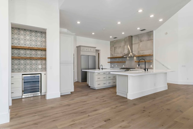 kitchen with light hardwood / wood-style floors, wall chimney exhaust hood, a center island with sink, and beverage cooler