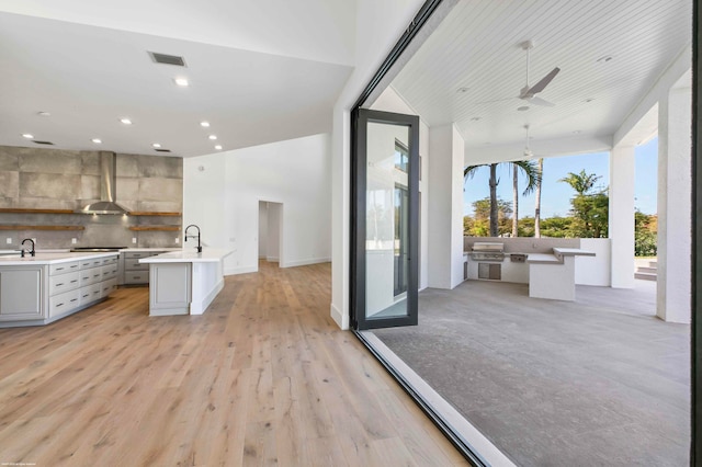 kitchen featuring wall chimney range hood, light wood-type flooring, an island with sink, gray cabinets, and decorative backsplash