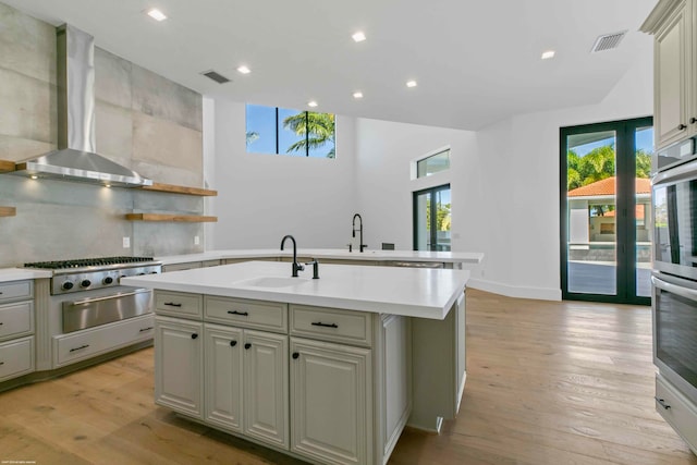 kitchen with wall chimney range hood, sink, light wood-type flooring, and an island with sink