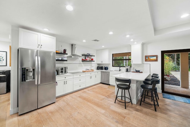 kitchen with a kitchen bar, appliances with stainless steel finishes, light wood-type flooring, wall chimney range hood, and white cabinets