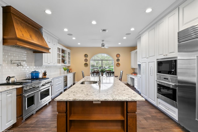 kitchen with built in appliances, white cabinets, custom range hood, a center island with sink, and light stone countertops