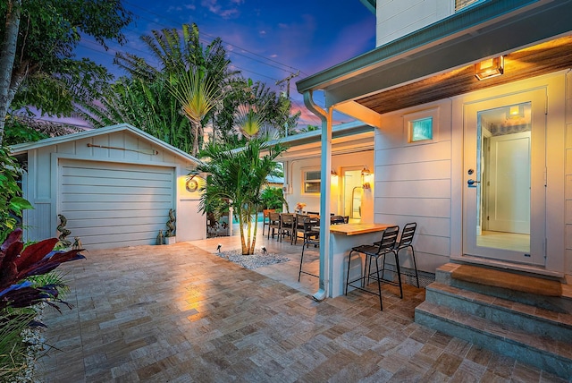 patio terrace at dusk featuring a garage, an outdoor structure, and an outdoor bar