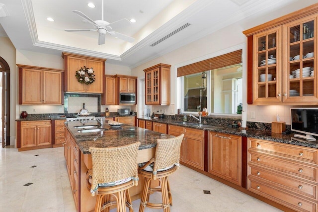 kitchen featuring a tray ceiling, ceiling fan, a center island with sink, and stainless steel microwave