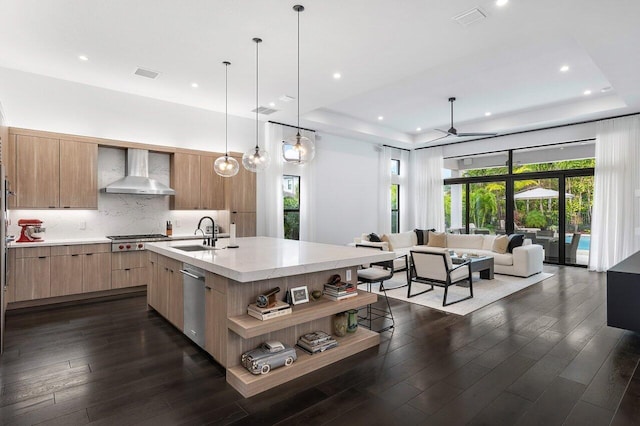 kitchen featuring appliances with stainless steel finishes, wall chimney exhaust hood, an island with sink, and a raised ceiling