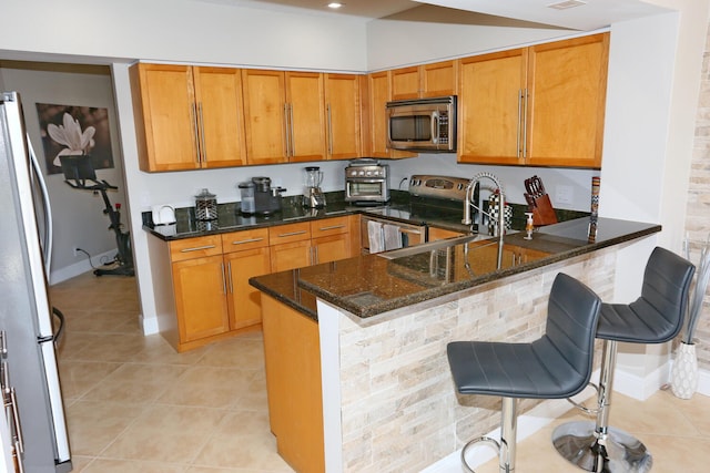 kitchen with kitchen peninsula, dark stone counters, stainless steel appliances, light tile flooring, and a kitchen bar