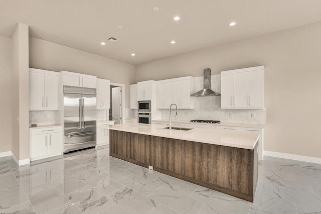 kitchen featuring wall chimney exhaust hood, sink, built in appliances, a center island with sink, and white cabinets