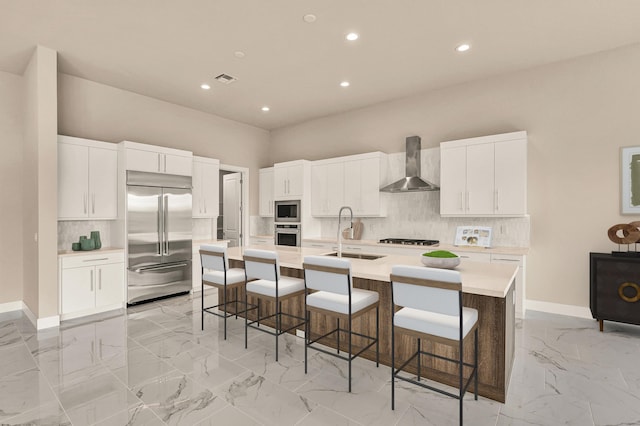 kitchen featuring sink, wall chimney range hood, built in appliances, an island with sink, and a kitchen bar