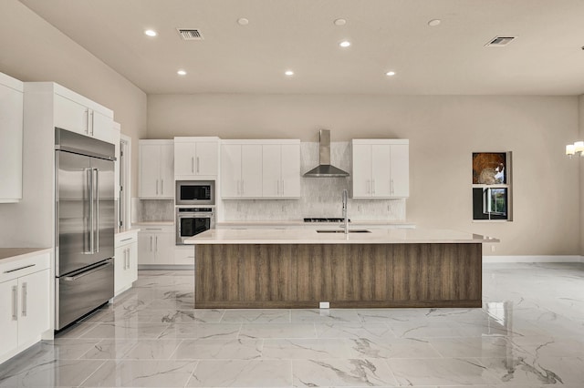 kitchen featuring built in appliances, wall chimney exhaust hood, white cabinets, and a kitchen island with sink