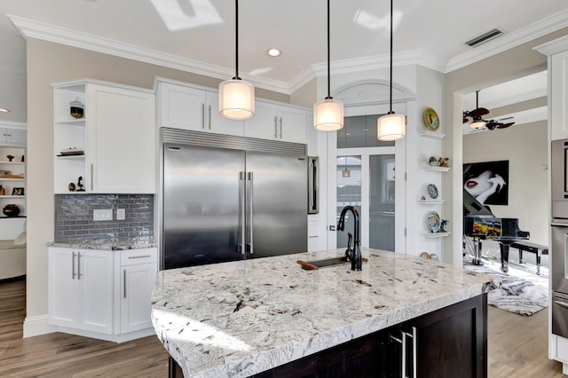 kitchen with stainless steel built in refrigerator, white cabinetry, light stone countertops, a center island with sink, and pendant lighting