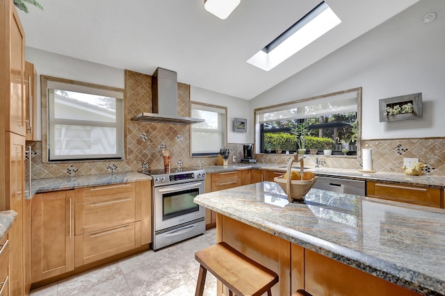 kitchen with light stone countertops, appliances with stainless steel finishes, decorative backsplash, vaulted ceiling with skylight, and wall chimney range hood