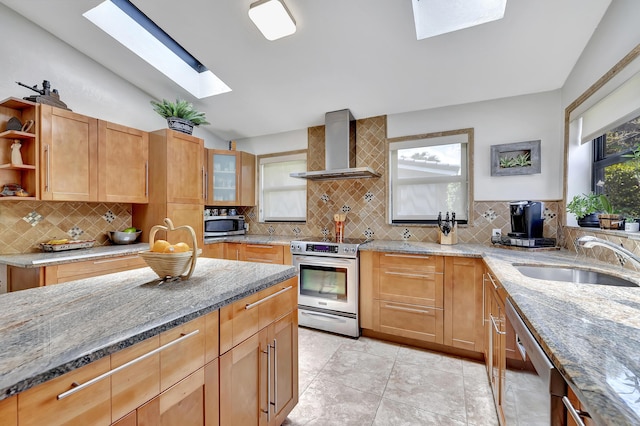 kitchen with vaulted ceiling with skylight, wall chimney exhaust hood, light stone counters, and appliances with stainless steel finishes