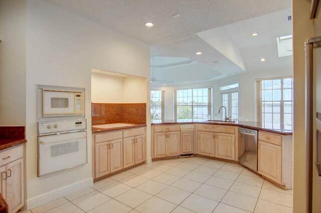 kitchen with a tray ceiling, white appliances, sink, light tile floors, and light brown cabinetry