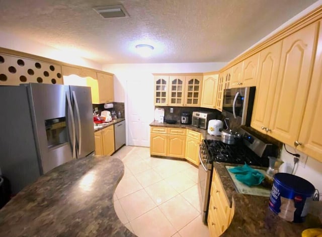 kitchen with light tile floors, appliances with stainless steel finishes, dark stone countertops, light brown cabinets, and a textured ceiling