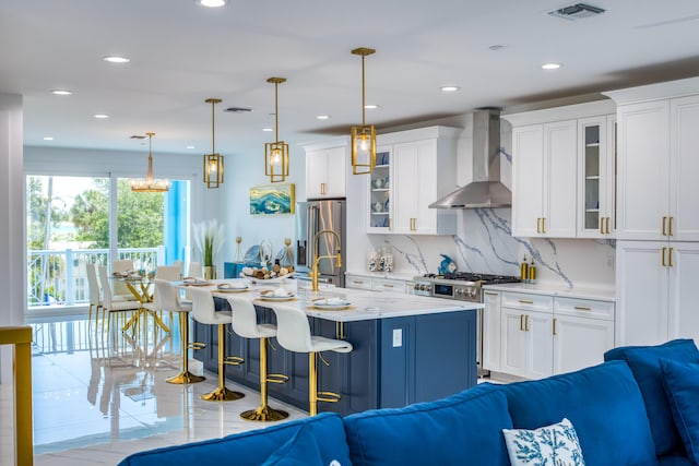 kitchen with hanging light fixtures, appliances with stainless steel finishes, white cabinets, wall chimney range hood, and a center island with sink