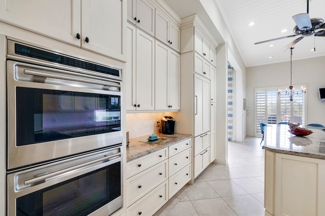 kitchen featuring double oven, light tile flooring, ceiling fan with notable chandelier, tasteful backsplash, and light stone counters