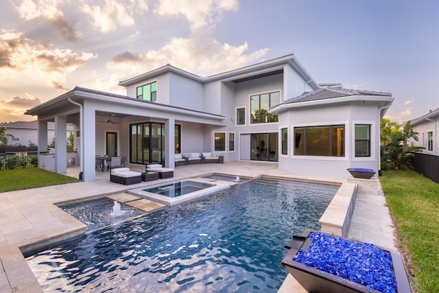 back house at dusk with an outdoor hangout area, ceiling fan, a swimming pool with hot tub, a patio area, and exterior kitchen