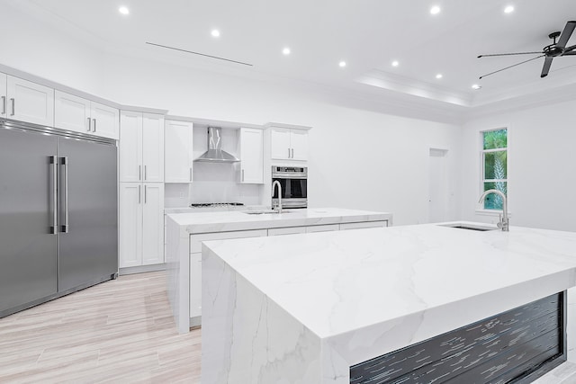 kitchen with a center island with sink, appliances with stainless steel finishes, sink, ceiling fan, and wall chimney range hood