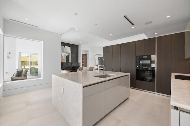 kitchen featuring light tile floors, a center island with sink, sink, black double oven, and light stone counters