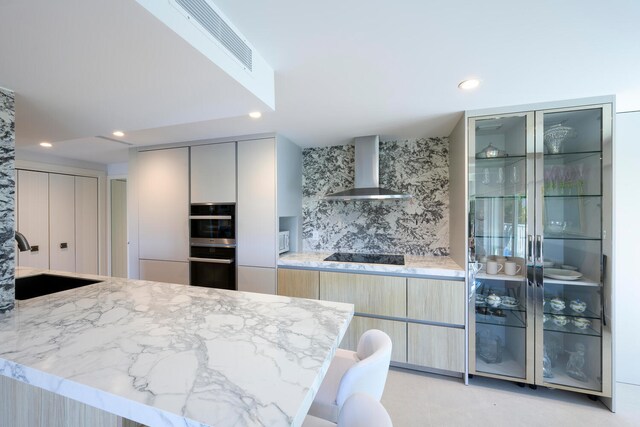 kitchen featuring sink, light stone counters, stainless steel double oven, a breakfast bar area, and wall chimney exhaust hood