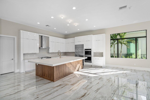 kitchen with sink, a kitchen island with sink, white cabinets, decorative backsplash, and wall chimney exhaust hood