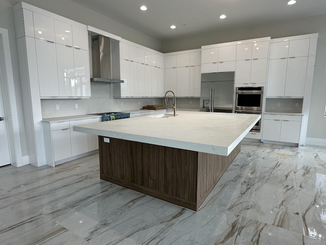 kitchen featuring sink, stainless steel appliances, wall chimney range hood, an island with sink, and white cabinets