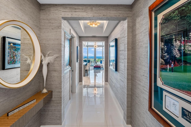 hall featuring a raised ceiling, tile walls, a notable chandelier, and light tile floors