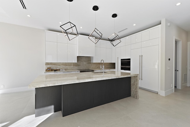 kitchen featuring decorative light fixtures, light stone countertops, a center island with sink, white cabinets, and backsplash