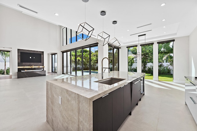 kitchen featuring a kitchen island with sink, sink, light tile floors, hanging light fixtures, and light stone countertops