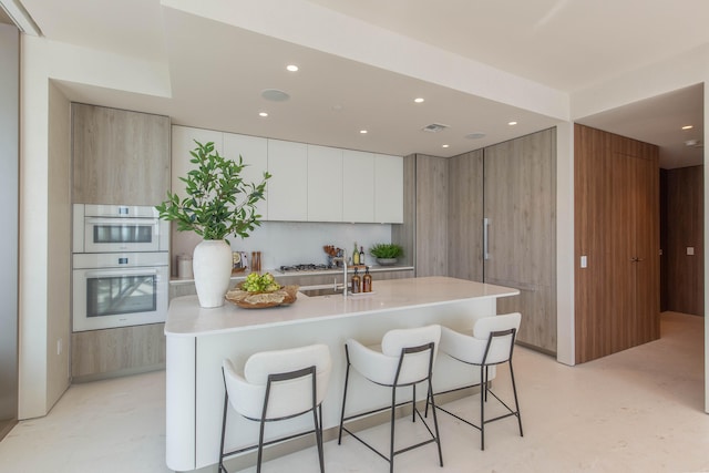 kitchen with double oven, a kitchen breakfast bar, stainless steel gas stovetop, white cabinetry, and a kitchen island with sink