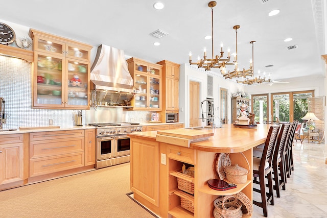 kitchen with wall chimney exhaust hood, decorative light fixtures, range with two ovens, backsplash, and an island with sink