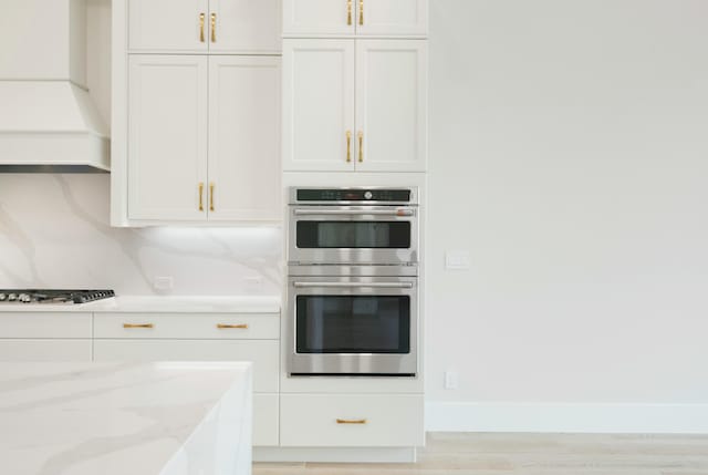 kitchen featuring light stone counters, custom exhaust hood, backsplash, white cabinets, and appliances with stainless steel finishes