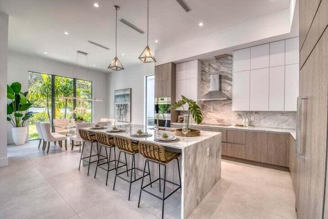 kitchen featuring an island with sink, white cabinets, hanging light fixtures, a kitchen breakfast bar, and wall chimney exhaust hood