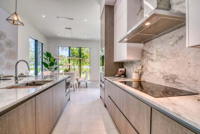 kitchen with black electric cooktop, pendant lighting, light brown cabinetry, sink, and wall chimney range hood