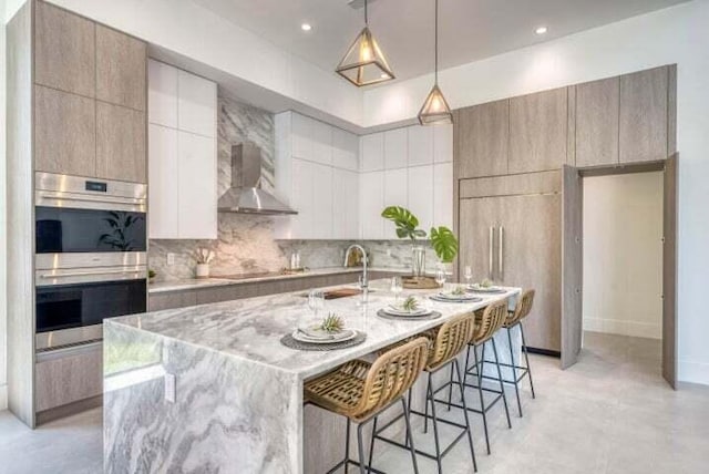 kitchen featuring pendant lighting, a breakfast bar area, an island with sink, wall chimney exhaust hood, and double oven