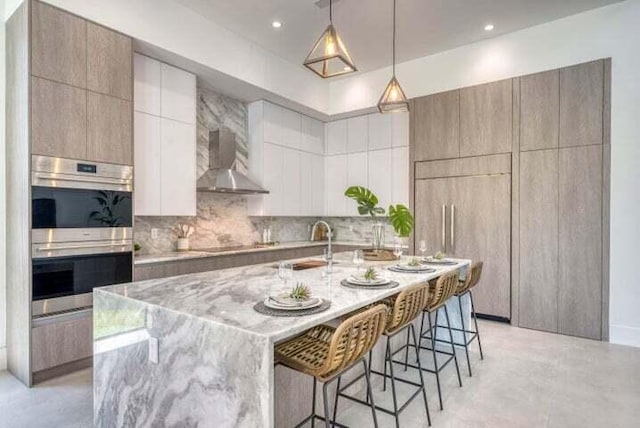 kitchen featuring a breakfast bar, a large island with sink, white cabinetry, wall chimney range hood, and double oven