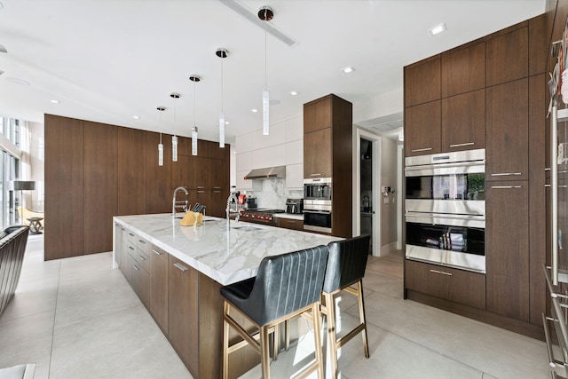 kitchen featuring light stone countertops, decorative light fixtures, stainless steel double oven, a breakfast bar, and a center island with sink