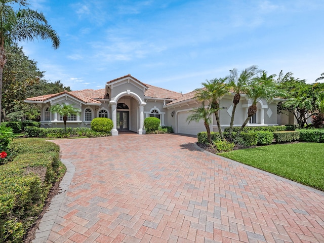 mediterranean / spanish house featuring a front lawn, a tile roof, stucco siding, decorative driveway, and an attached garage