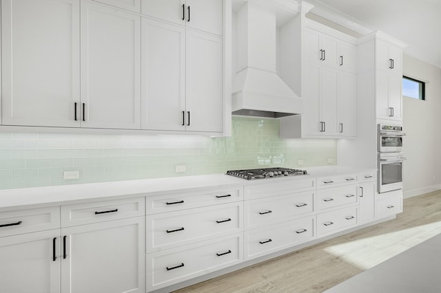 kitchen with custom exhaust hood, stainless steel appliances, decorative backsplash, white cabinets, and light hardwood / wood-style flooring