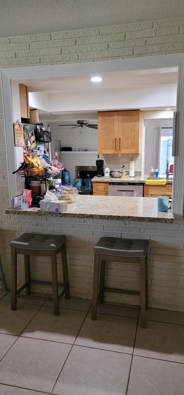 kitchen with light brown cabinetry, a breakfast bar area, light stone counters, dishwasher, and a textured ceiling