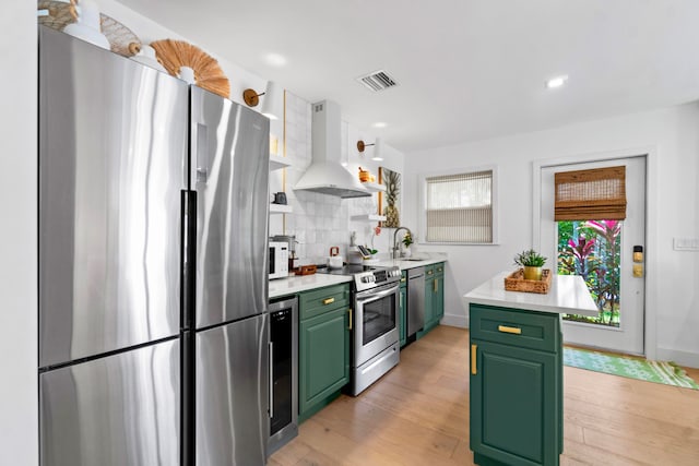 kitchen featuring stainless steel appliances, wall chimney range hood, green cabinets, light hardwood / wood-style flooring, and backsplash