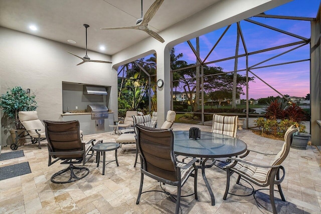 patio terrace at dusk with grilling area, a lanai, an outdoor kitchen, and ceiling fan