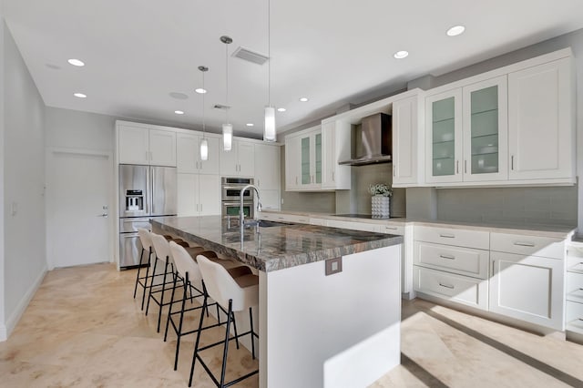 kitchen with appliances with stainless steel finishes, wall chimney exhaust hood, a kitchen island with sink, pendant lighting, and white cabinets