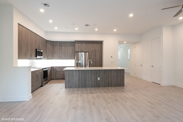 kitchen featuring a center island with sink, sink, ceiling fan, light wood-type flooring, and appliances with stainless steel finishes