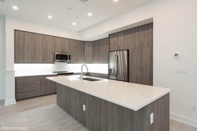 kitchen featuring a kitchen island with sink, sink, appliances with stainless steel finishes, light hardwood / wood-style floors, and light stone counters